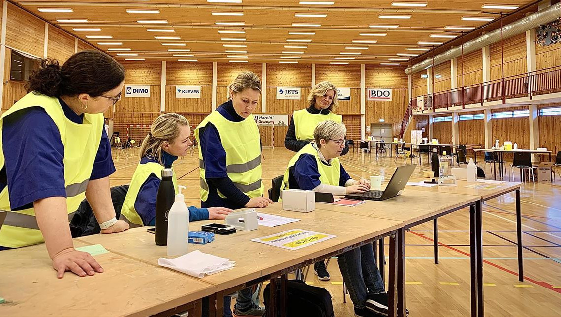 personar i ein idrettshall som lener seg over eit bord med datamaskin. Personane har på seg refleksvest