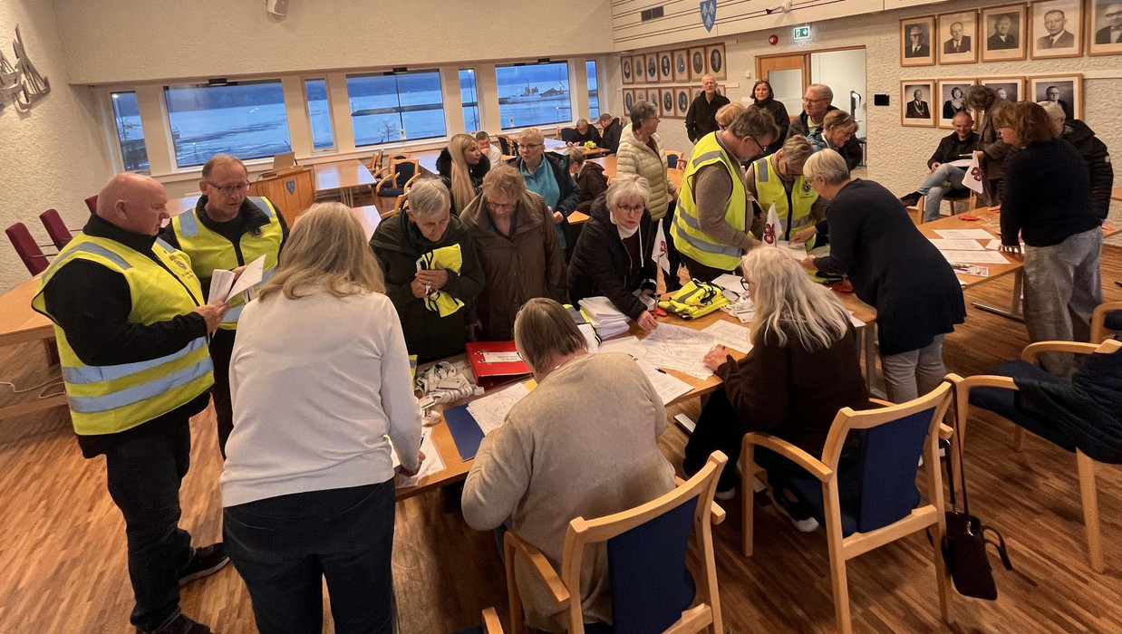 mange personar rundt som står rundt forskjellige bord og ser på papir, skjema og brosjyrer. Mange har gul refleksvest på seg. I kommunestyresalen.